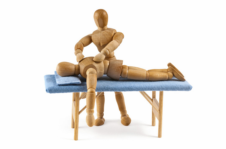 Two wooden artist's mannequins: one kneeling behind another lying face-down on a small folding massage couch with a blue towel