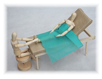 A wooden mannequin on a stool working on the feet of a second mannequin reclined under a sheet of green couch roll